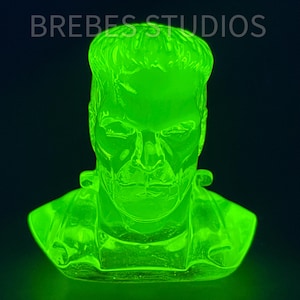 May include: A neon green bust of Frankenstein's monster. The sculpture is lit from within, creating a glowing effect. The bust is a detailed representation of the classic movie character. The text "BREBES STUDIOS" is at the top.