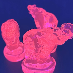 May include: Three pink, translucent sculptures of elephant heads on a dark blue background. The sculptures are lit with a pink glow, highlighting their form and texture. The elephant heads are on a round base.