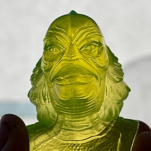 May include: A translucent, lime-green figurine depicting a creature with a scaled, amphibious head, prominent brow ridges, wide eyes, and a textured neck. The sculpture features detailed facial contours and gill-like structures on the sides.