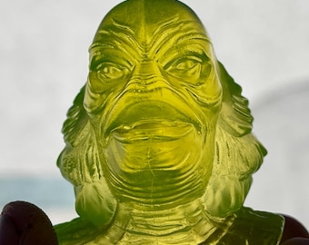May include: A translucent, lime-green figurine depicting a creature with a scaled, amphibious head, prominent brow ridges, wide eyes, and a textured neck. The sculpture features detailed facial contours and gill-like structures on the sides.