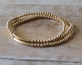 May include: A set of two gold-toned beaded bracelets featuring small, round, reflective metallic beads and a single smooth, curved, cylindrical gold-toned tube bead integrated into each strand, creating a uniform and polished appearance.