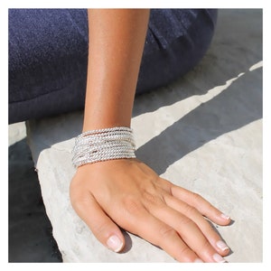 May include: A silver beaded bracelet stacked on a person's wrist. The bracelet is made of small, round beads that are tightly woven together.