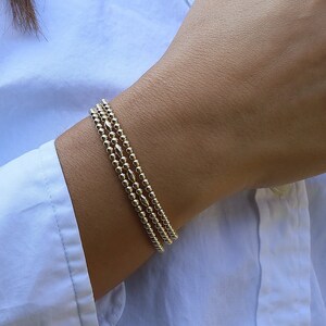 May include: A set of three gold-toned beaded bracelets featuring small, uniform spherical beads and occasional elongated, diamond-shaped accent beads. The bracelets are worn together, creating a layered appearance with a metallic, reflective finish.