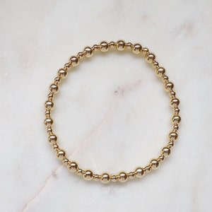 May include: A gold beaded bracelet featuring small, round beads of varying sizes. The beads are strung together to create a circular design. The bracelet is presented on a white marble surface.