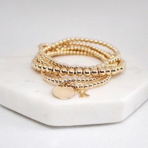 Könnte beinhalten: Ein Set aus vier goldenen Perlenarmbändern mit einem kleinen goldenen Anhänger. Der Anhänger trägt den Buchstaben "K".