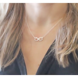 May include: A silver infinity necklace worn by a model with blonde hair. The necklace is simple and delicate, featuring a small infinity symbol pendant.