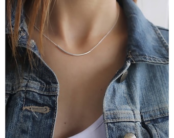 May include: A silver-toned necklace featuring a thin, curved metal bar pendant centered on a delicate chain. The necklace is worn against a white top layered under a blue denim jacket with visible metal buttons, one of which is embossed with the text gap.