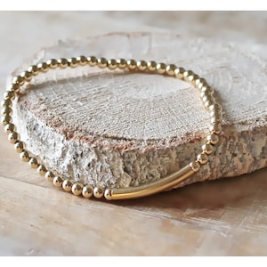 Op de afbeelding: Een goudkleurige kralenarmband bestaande uit een reeks kleine, uniforme bolvormige kralen afgewisseld met een enkele, gladde, langwerpige cilindrische staafkraal. De armband is cirkelvormig en heeft een gepolijste, reflecterende metalen afwerking.