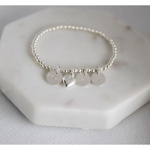 Peut inclure: Un bracelet élastique composé de perles argentées avec quatre breloques suspendues : un disque circulaire gravé du mot MOM, un cœur bombé lisse et deux autres disques circulaires gravés des lettres J et K, le tout en finition métallique.