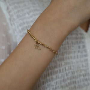 May include: Gold beaded bracelet with a dangling letter "M" charm. The bracelet is made of small, round gold beads and is worn on a wrist. The charm is a cursive letter "M".