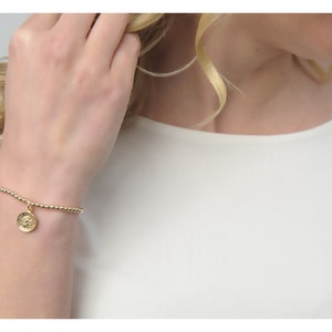 Könnte beinhalten: Goldenes Perlenarmband mit rundem Anhänger. Der Anhänger hat ein detailliertes Design. Das Armband wird am Handgelenk getragen, mit einem weißen Oberteil im Hintergrund. Es ist ein zarter Schmuck.