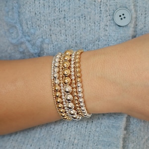 May include: A stack of five beaded bracelets featuring alternating gold and silver metallic spheres. The bracelets vary in bead size, with some strands consisting of small, uniform metallic balls and others featuring larger, polished gold and silver beads.