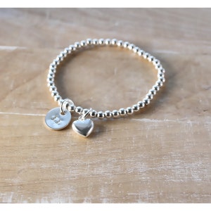 Könnte beinhalten: Ein silbernes Armband mit Perlen, einem kleinen Herz-Anhänger und einem runden Anhänger mit dem eingravierten Buchstaben "B".
