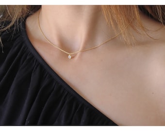 May include: A delicate gold chain necklace featuring a single, small, round, clear gemstone pendant centered on the collarbone. The necklace is worn against a black garment, highlighting the contrast between the metallic gold and the stone.