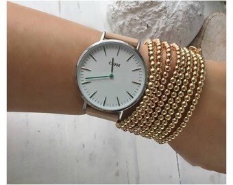 May include: A wristwatch with a silver-tone round case, a white dial featuring the text CLUSE, black hour markers, and a green second hand, paired with a beige leather strap and a stack of multiple gold-tone beaded stretch bracelets.