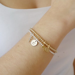 May include: Two beaded stretch bracelets, one in silver and one in gold, featuring small dangling charms. The silver bracelet includes a round disc charm engraved with the letter C and a cross charm. The gold bracelet features a cross and a disc charm.