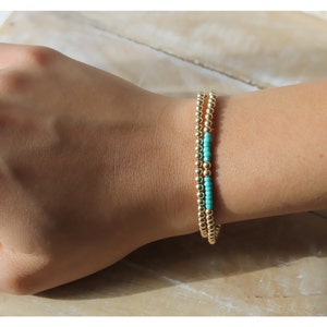 Op de afbeelding: Twee gouden kralen armbanden gestapeld op een pols. De bovenste armband heeft een enkele rij turquoise kralen.