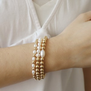 May include: Three gold bead bracelets with white pearl accents. The bracelets are stacked on a person's wrist.
