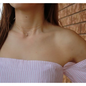May include: A woman wearing a pink and white striped off-the-shoulder top with a gold chain necklace featuring a small cross pendant.