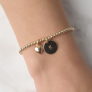May include: Gold beaded bracelet with a heart and a round charm with the letter K engraved on it.
