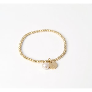 Könnte beinhalten: Ein goldfarbenes Armband mit einer einzelnen Perle und einem goldenen Anhänger mit der eingravierten Zahl "1".