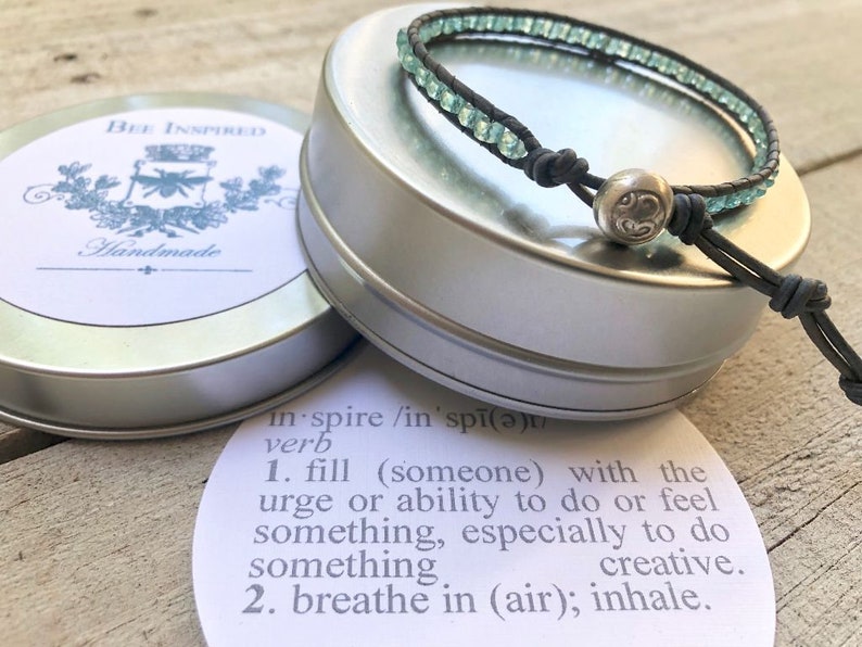 May include: A wrap bracelet with light blue beads and a silver clasp rests on a silver tin. The bracelet is made of black leather. The tin has a label that says "BEE INSPIRED Handmade". A definition of the word "inspire" is also visible.