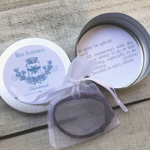 May include: A small, round, silver tin with the words "BEE INSPIRED" and "Handmade" printed on the lid. Inside, a bracelet is visible in a sheer white bag. The tin's interior features a definition of "inspire".