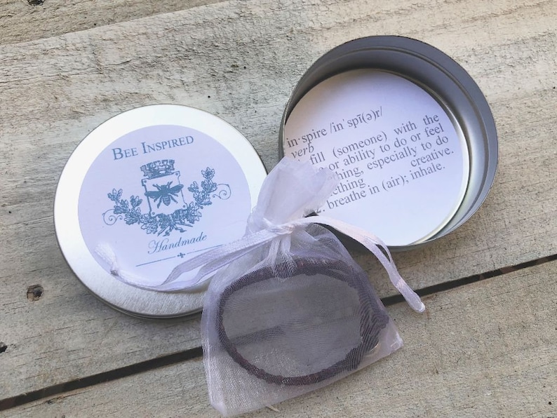May include: A small, round, silver tin with the words "BEE INSPIRED" and "Handmade" printed on the lid. Inside, a bracelet is visible in a sheer white bag. The tin's interior features a definition of "inspire".