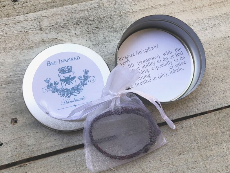 Op de afbeelding: Een kleine, ronde, zilveren blik met een wit label waarop "BEE INSPIRED" en "Handmade" staat. De blik is open en een klein, bruin armbandje zit in een wit organza zakje.