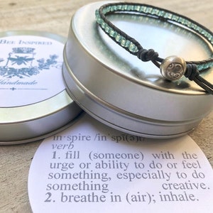 Op de afbeelding: Een turquoise en zilveren armband met een leren koord en een zilveren bedel. De armband ligt op een witte achtergrond met een definitie van het woord "inspireren" op een kaart.