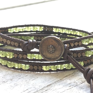 Op de afbeelding: Een bruin leren wikkelarmband met groene edelsteen kralen en een messing knoopsluiting.