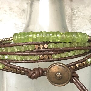Op de afbeelding: Een multi-wrap armband met bruin lederen koord, groene edelsteen kralen en goudkleurige metalen accenten. De armband heeft een grote, ronde, goudkleurige metalen sluiting.