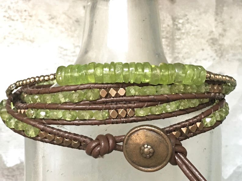 Op de afbeelding: Een multi-wrap armband met bruin lederen koord, groene edelsteen kralen en goudkleurige metalen accenten. De armband heeft een grote, ronde, goudkleurige metalen sluiting.