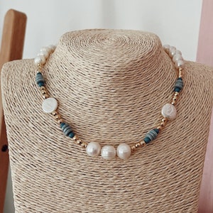 Könnte beinhalten: Eine weiße Perlen- und Goldkette mit blaugrünen und weißen Perlen. Die Kette befindet sich auf einer strohfarbenen Puppe.