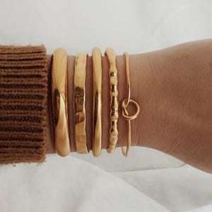 Pode incluir: Uma coleção de pulseiras e braceletes dourados. As pulseiras variam em design, incluindo estilos lisos, texturizados e em forma de nó. As joias são usadas em um pulso, com uma manga de suéter marrom visível no fundo.
