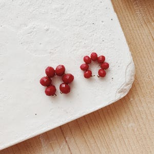 Può includere: Due orecchini a forma di cerchio con un'apertura al centro, realizzati con perline di vetro rosso. Le perline sono sfaccettate e hanno una finitura lucida.