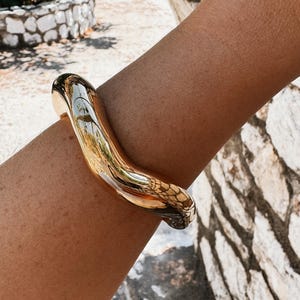 Pode incluir: Uma pulseira dourada brilhante, com uma forma curva e orgânica. A pulseira grossa e maciça reflete o ambiente, criando um efeito espelhado. A joia é usada no braço.