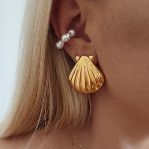 Op de afbeelding: Een gouden schelpvormige oorbel en een witte parelmanchet worden op een oor getoond. De schelpoorbellen hebben een gedetailleerd, gestructureerd ontwerp. De oormanchet heeft drie witte parels.