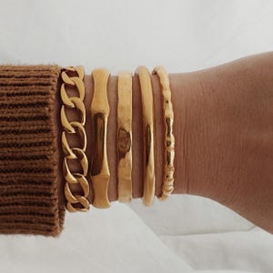 May include: A set of five gold-toned bracelets, including a chain link bracelet and four bangle bracelets. The bangles are different widths and have various textures.