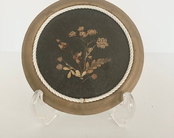 May include: A round, wooden plate with a white rope border. The plate has a dark background with a pressed flower design in the center.