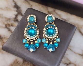 Puede incluir: Un par de pendientes colgantes con acabado dorado. Los pendientes presentan una gran cuenta azul turquesa rodeada de cuentas más pequeñas de color azul turquesa y blanco. Los pendientes tienen un diseño en cascada con múltiples capas de cuentas.