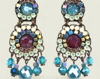Puede incluir: Un par de pendientes ornamentados con una estética vintage. Cada pendiente presenta una gema grande, facetada de color turquesa en la parte inferior, con gemas y cuentas turquesas y azules más pequeñas. Los pendientes tienen una gema central granate.