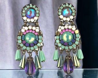 以下が含まれることがあります： マルチカラーのデザインのドロップイヤリングのペア。イヤリングは、虹色の緑色の丸い中心に、虹色の緑色、青色、ピンク色の小さなビーズが囲まれています。イヤリングは、紫と緑のクリスタル製のドロップで飾られています。