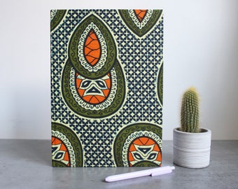 May include: A rectangular notebook with a colorful pattern featuring orange, green, and navy blue geometric shapes. A small cactus in a gray pot and a white pen are next to the notebook on a gray surface.