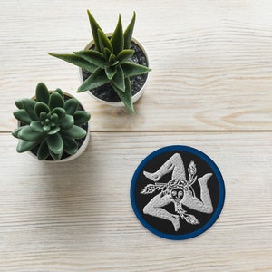 May include: A white and silver embroidered patch of the Trinacria symbol, a three-legged woman holding a head of wheat in each hand, on a black background with a blue border. The patch is on a light wood surface with two small green succulent plants in white pots.