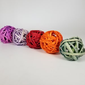 May include: Five spherical decorative balls made of woven rattan strips. The balls are colored in shades of deep purple, light lavender, vibrant red, bright orange, and muted sage green, arranged in a diagonal line against a plain white background.