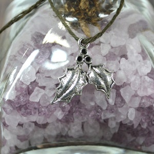 Op de afbeelding: Een glazen fles gevuld met paarse badzout en een zilveren hulstbladhanger die aan de bovenkant hangt.