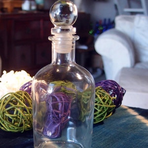 Pode incluir: Uma garrafa de vidro transparente com uma rolha redonda de vidro. A garrafa está vazia e tem uma superfície texturizada. A garrafa está sobre uma superfície com um padrão azul e branco.