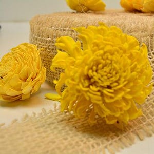 May include: Bright yellow artificial flowers with layered petals arranged on a roll of natural tan burlap ribbon. The textured fabric ribbon features a frayed edge and is coiled, providing a base for the vibrant yellow floral accents.