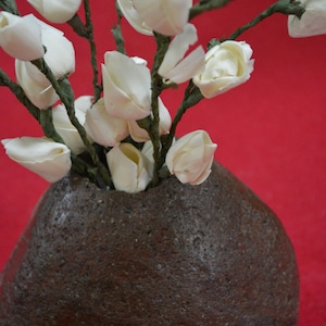 Puede incluir: Un ramo de flores de papel blanco en un jarrón de cerámica marrón oscuro texturizado. El jarrón tiene la forma de una roca redondeada.